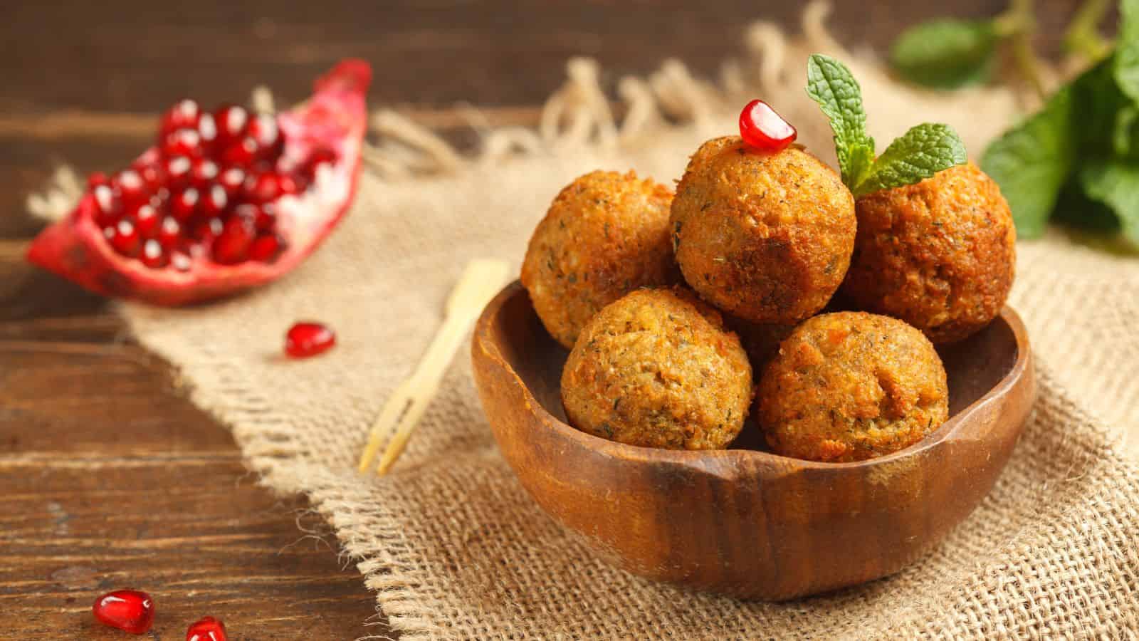 A wooden bowl filled with five falafel balls garnished with a mint sprig and a pomegranate seed, placed on a piece of burlap. There is a halved pomegranate and scattered seeds in the background.