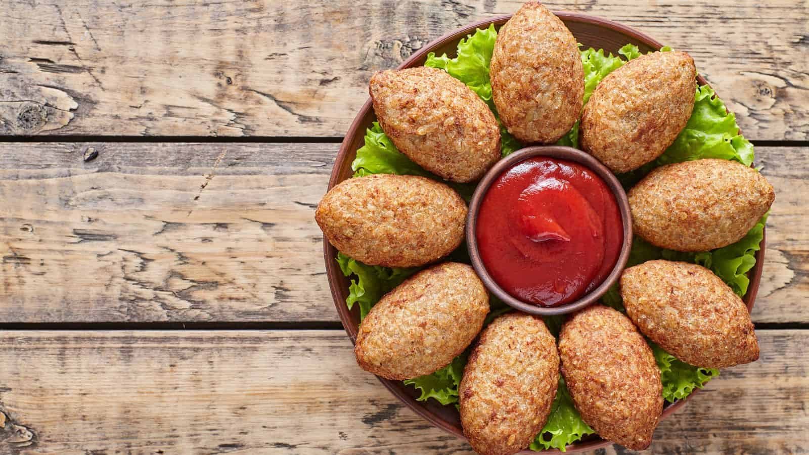 A round wooden plate holds nine kibbeh arranged in a circle on a bed of lettuce around a small bowl of red sauce, all set on a rustic wooden table.
