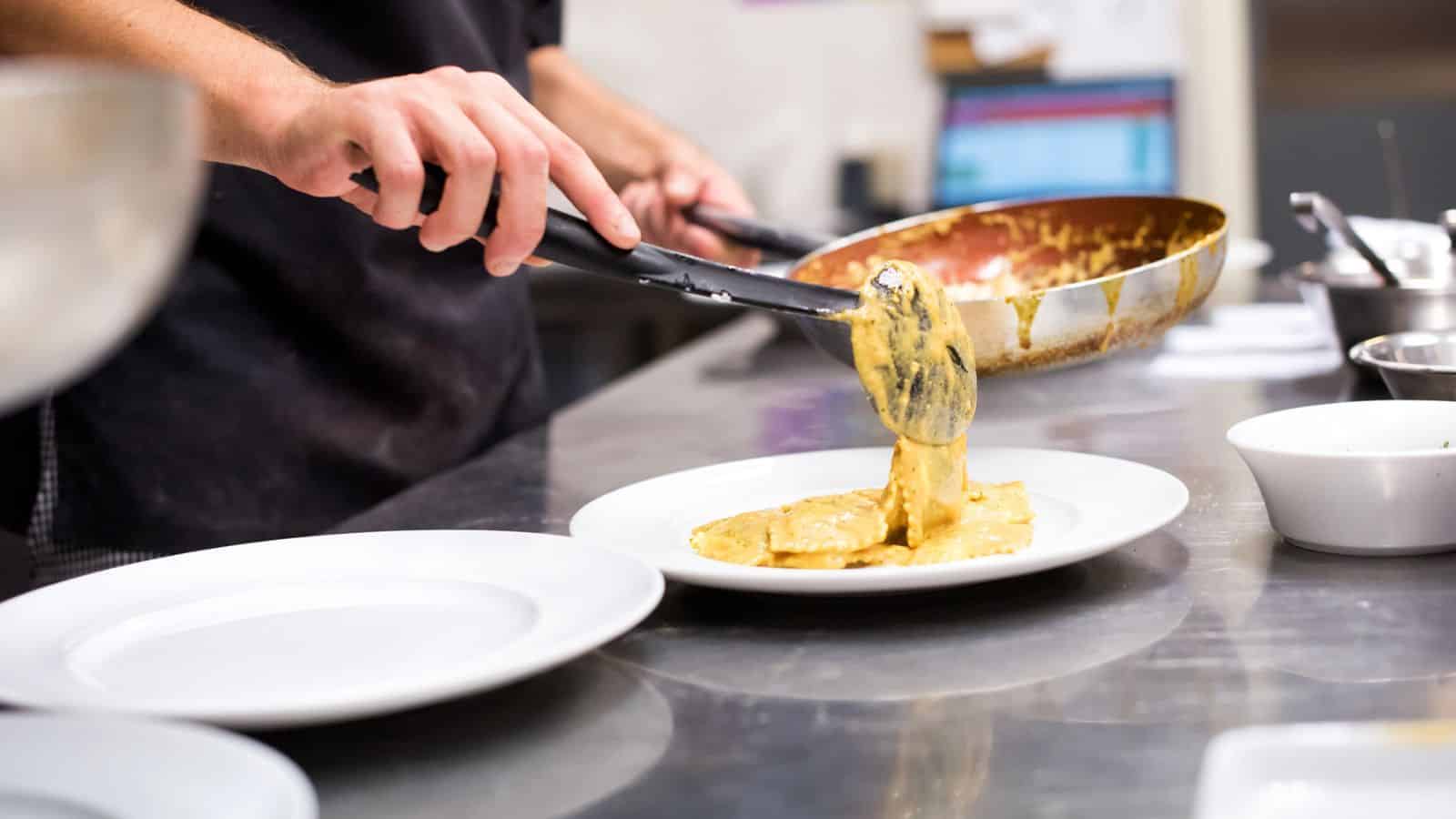 A person uses tongs to plate pasta in a creamy yellow sauce from a pan onto a white plate in a professional kitchen.