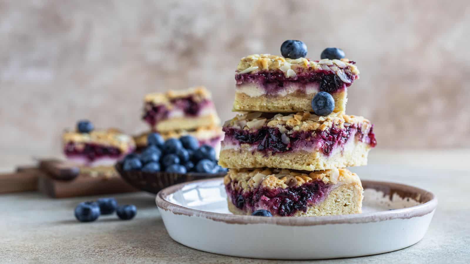 Three stacked dessert bars with a crumbly crust, creamy filling, and blueberry topping are on a plate, with fresh blueberries scattered around and more bars in the background.