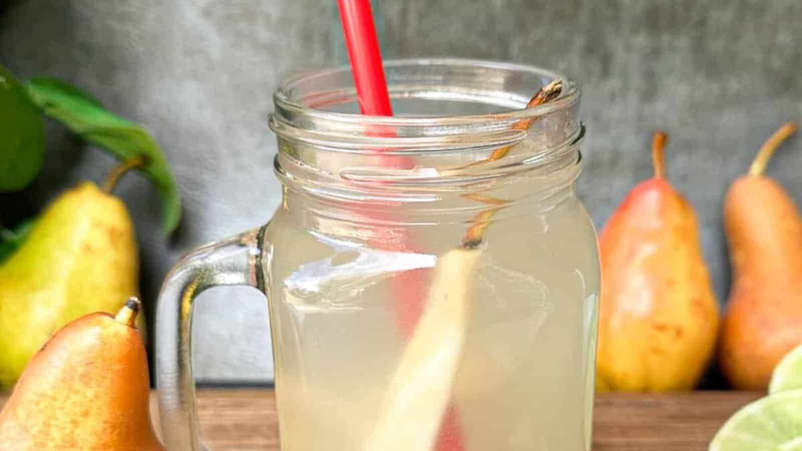 A glass mug filled with pear tea juice and a red straw, surrounded by whole pears, lime slices, and ginger on a wooden surface.