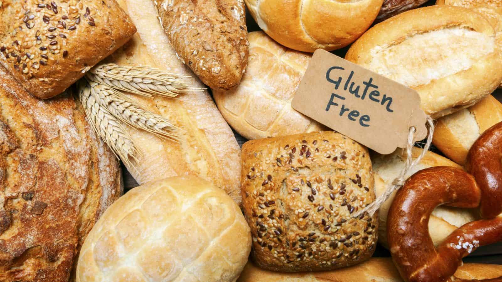 Assorted loaves and rolls of bread, including seeded and plain varieties, are arranged next to a small sign labeled “Gluten Free.” Wheat stalks are also visible among the baked goods.