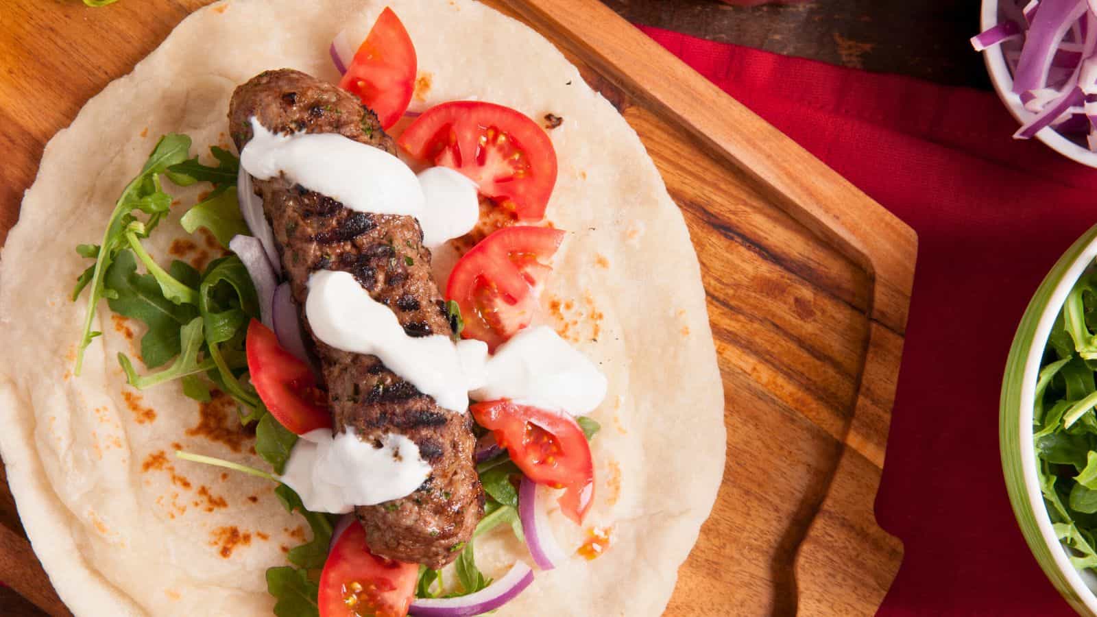 A grilled beef kebab on flatbread is topped with sliced tomatoes, red onions, arugula, and white sauce, served on a wooden board with a bowl of greens and red onions beside it.