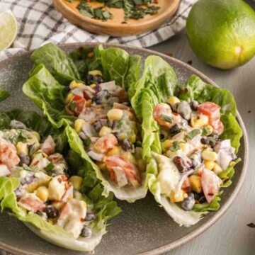 Three romaine lettuce leaves filled with a mixture of corn, black beans, diced tomatoes, and creamy dressing sit on a plate. Nearby are a halved avocado, a bowl of chili flakes, limes, and a checkered cloth.