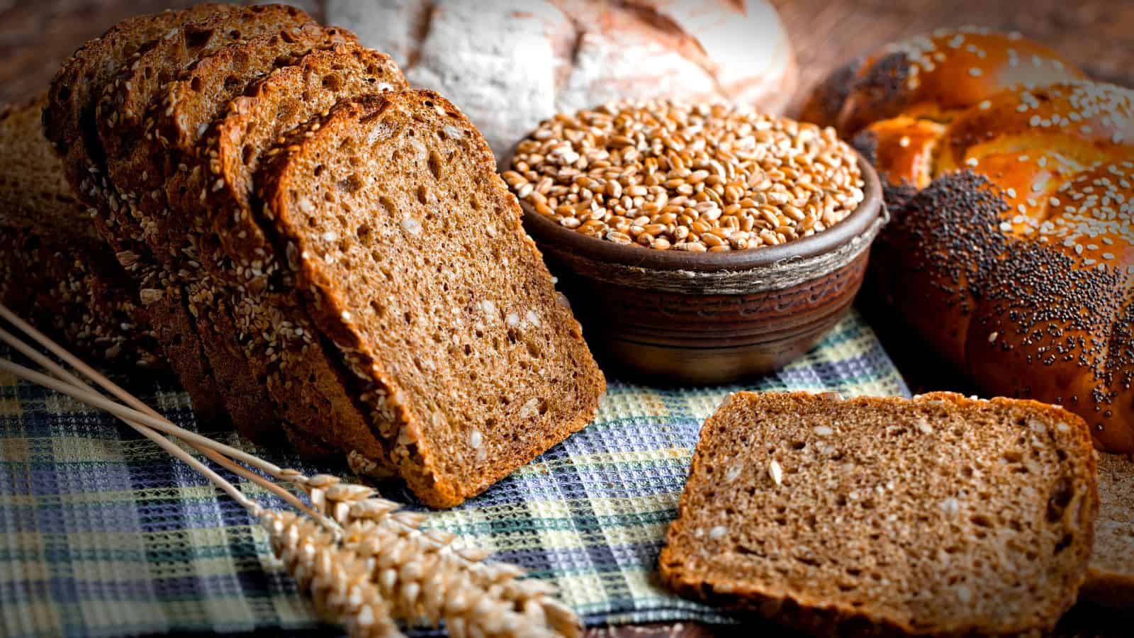 Several slices of whole grain bread, a bowl of wheat grains, a wheat stalk, and other types of bread are arranged on a checkered cloth.