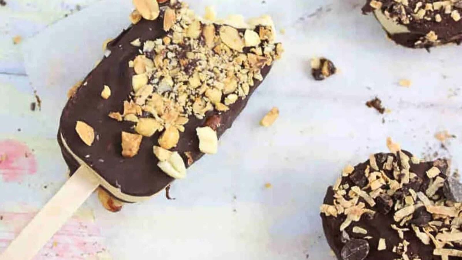 Homemade ice cream bar dipped in chocolate on a plate being covered in peanuts with more ice cream bars around it.