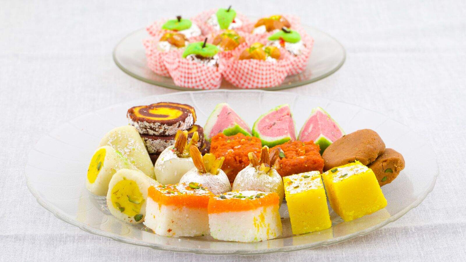 A glass plate with assorted Indian sweets, including barfi, peda, and rolled sweets, is in the foreground. In the background, another plate holds green and orange sweets in cupcake liners. Both plates are on a light surface.
