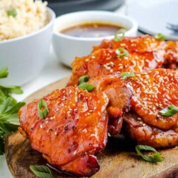 Three pieces of glazed chicken garnished with sesame seeds and chopped green onions sit on a wooden board, accompanied by fresh herbs, a bowl of rice, and a small container of sauce in the background.