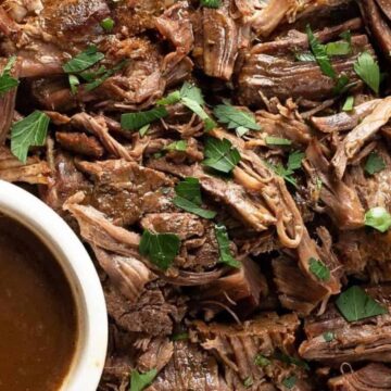 Shredded pot roast garnished with chopped fresh herbs, served alongside a small bowl of brown sauce or gravy on a white plate.