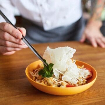 A person in a chef's coat holds chopsticks and places herbs on a bowl of noodles with vegetables and a crispy garnish, set on a wooden countertop.