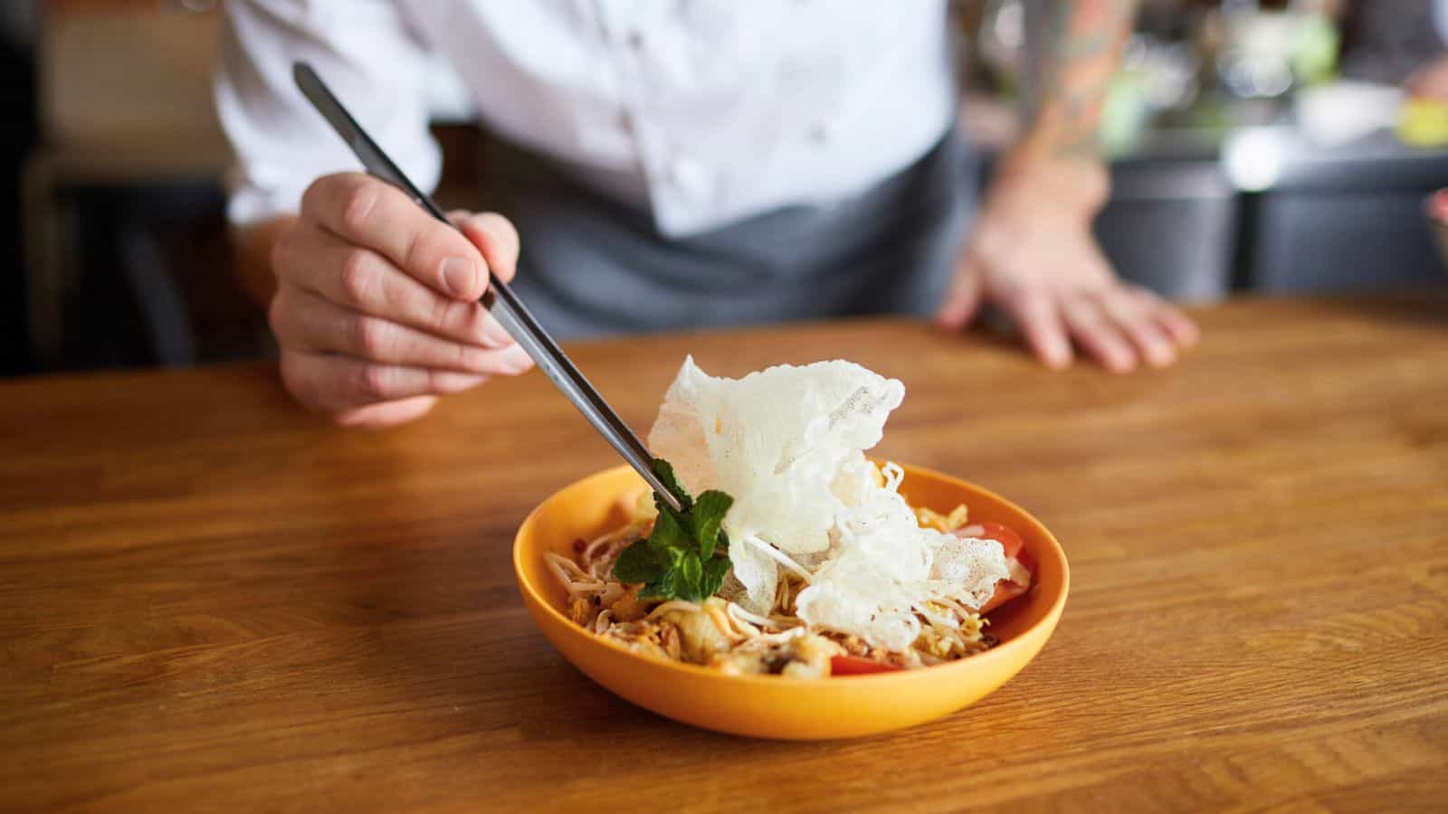 A person in a chef's coat holds chopsticks and places herbs on a bowl of noodles with vegetables and a crispy garnish, set on a wooden countertop.