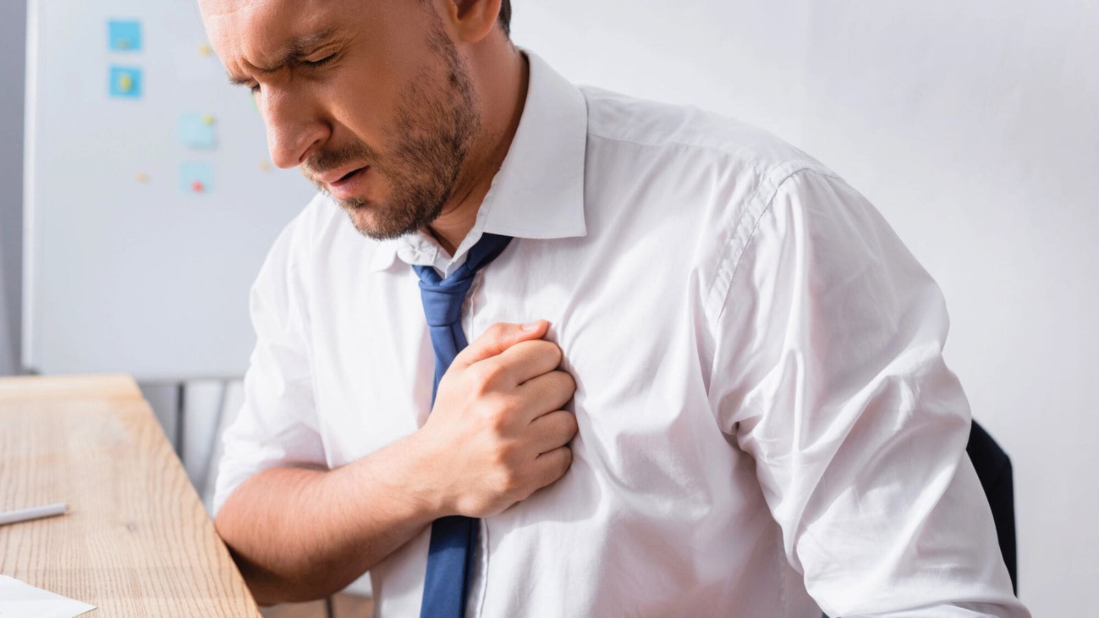 A man in a white shirt and tie sits at a desk, clutching his chest with a pained expression, suggesting discomfort or possible chest pain.