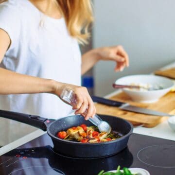 A woman in a white t-shirt is standing at a stove, seasoning a pan of vegetables. There are bowls and chopped ingredients on the countertop nearby.