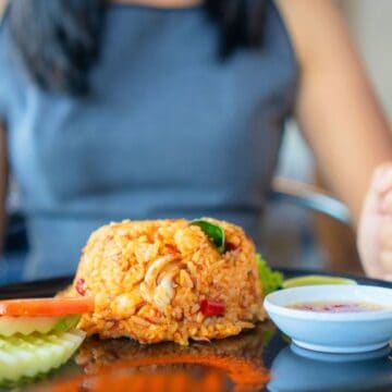 A person holding a fork and spoon sits in front of a plate of fried rice with sliced cucumbers, tomato, and a small dish of sauce. The person wears a sleeveless gray top.