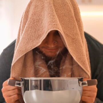 A person sits with a towel draped over their head, leaning over a steaming pot while holding it with both hands, in a kitchen setting.