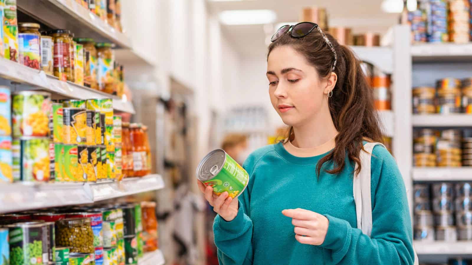 A woman wearing a green sweater stands in a grocery store aisle, holding and examining a can of food while surrounded by shelves stocked with canned goods and packaged products.