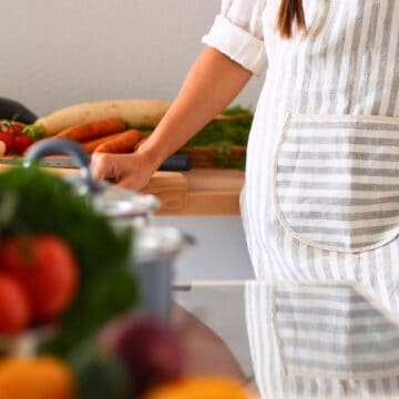 A person wearing a striped apron stands at a kitchen counter with various vegetables, including cauliflower, carrots, and tomatoes, visible on the countertop and in the foreground.