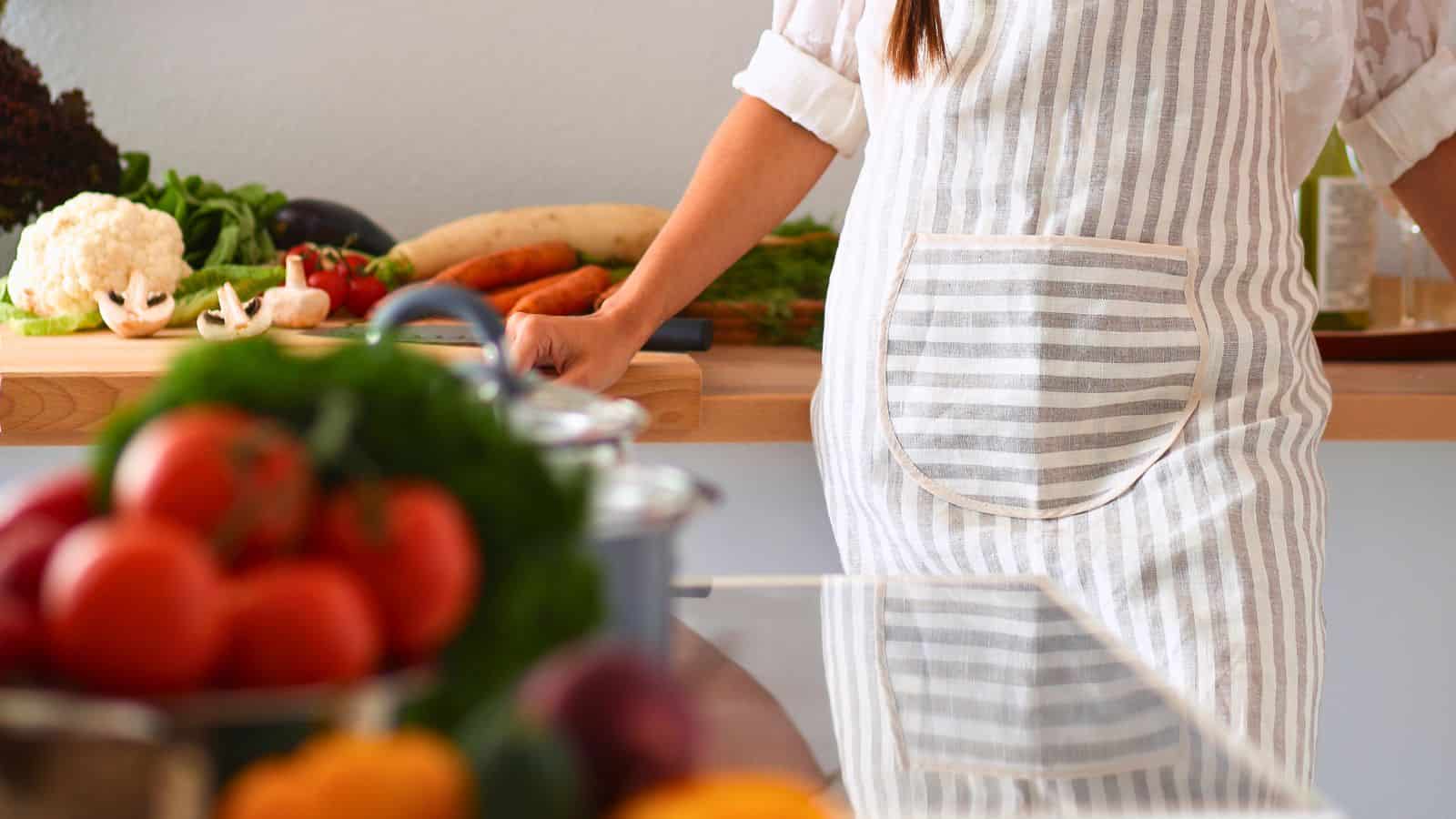 A person wearing a striped apron stands at a kitchen counter with various vegetables, including cauliflower, carrots, and tomatoes, visible on the countertop and in the foreground.