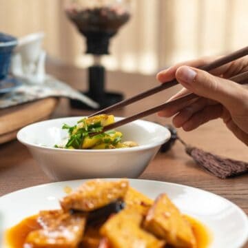 A person uses chopsticks to pick up food from a white bowl at a wooden table set with dishes, including a plate of tofu in sauce and traditional teaware in the background.