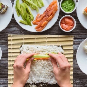A person prepares sushi by placing an avocado slice on rice and seaweed. Surrounding dishes hold sushi rolls, sliced fish, avocado, wasabi, ginger, rice, and cream cheese on a wooden table with a bamboo mat.