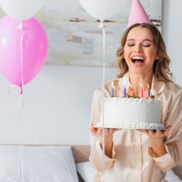 A woman wearing a pink party hat and pajamas is smiling and holding a birthday cake with lit candles, standing in a decorated room with white and pink balloons.