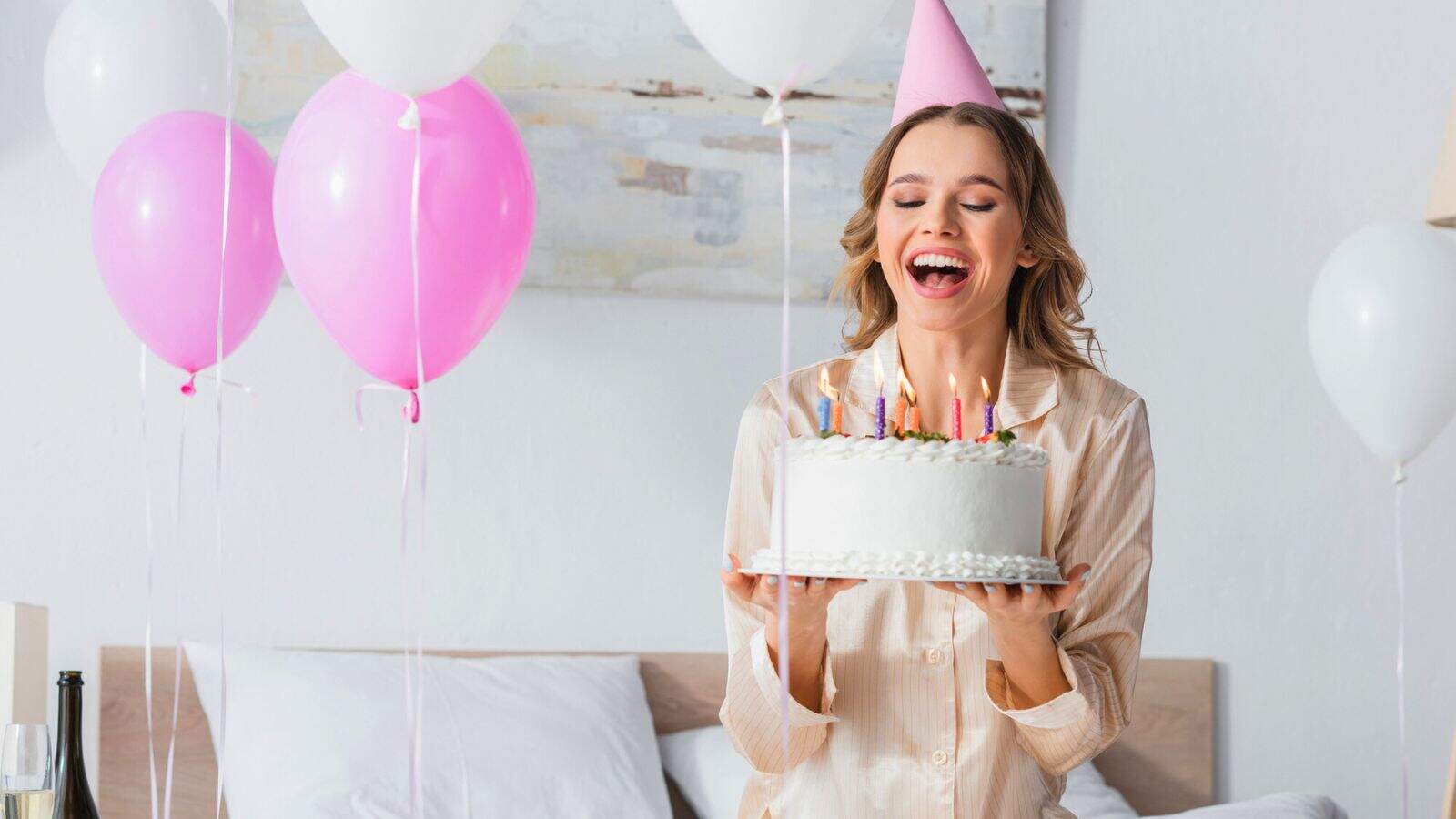 A woman wearing a pink party hat and pajamas is smiling and holding a birthday cake with lit candles, standing in a decorated room with white and pink balloons.