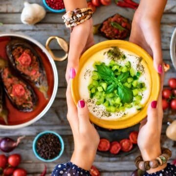 Two hands pass a bowl of yogurt dip topped with diced cucumber and herbs over a table with Mediterranean dishes, including stuffed eggplants, tomato-based stew, vegetables, spices, and kitchen utensils.