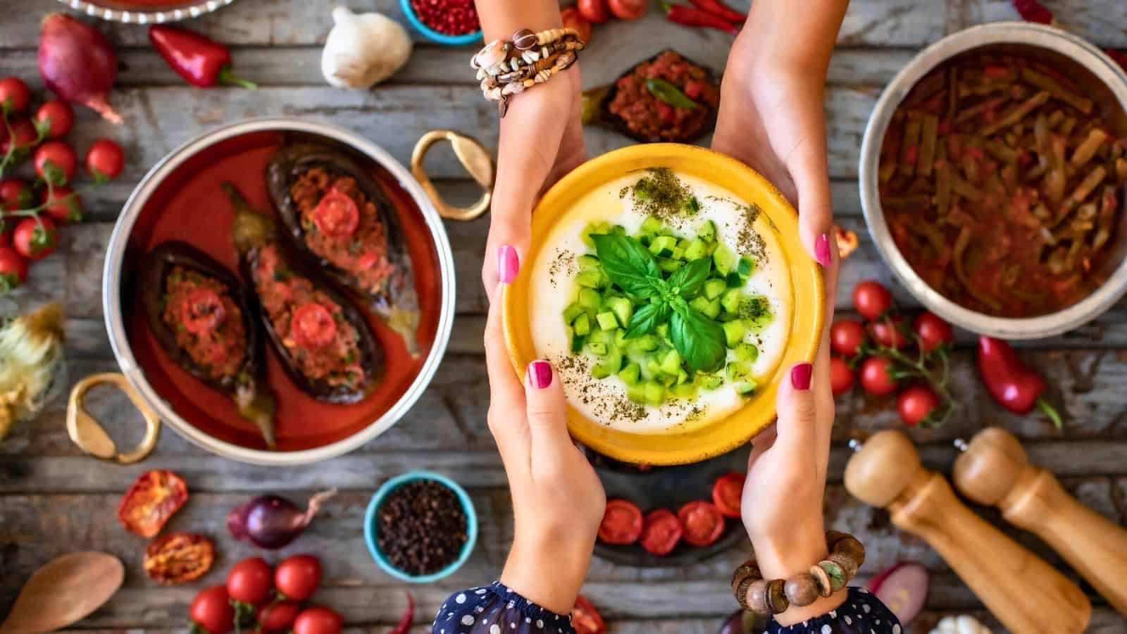 Two hands pass a bowl of yogurt dip topped with diced cucumber and herbs over a table with Mediterranean dishes, including stuffed eggplants, tomato-based stew, vegetables, spices, and kitchen utensils.