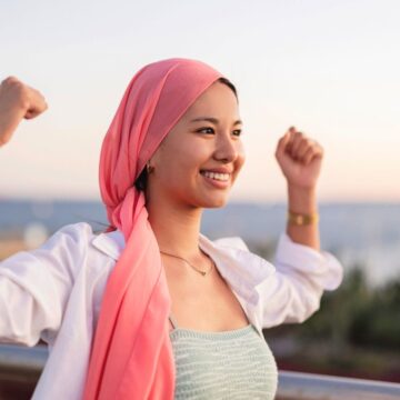 A woman wearing a pink headscarf and white shirt smiles and raises her arms in a confident pose outdoors, with blurred greenery and water in the background.