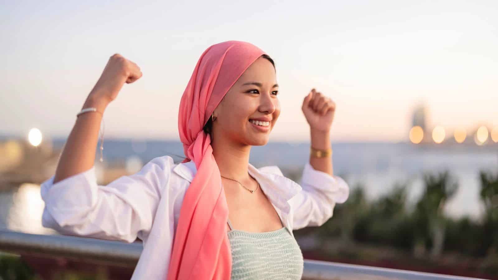 A woman wearing a pink headscarf and white shirt smiles and raises her arms in a confident pose outdoors, with blurred greenery and water in the background.