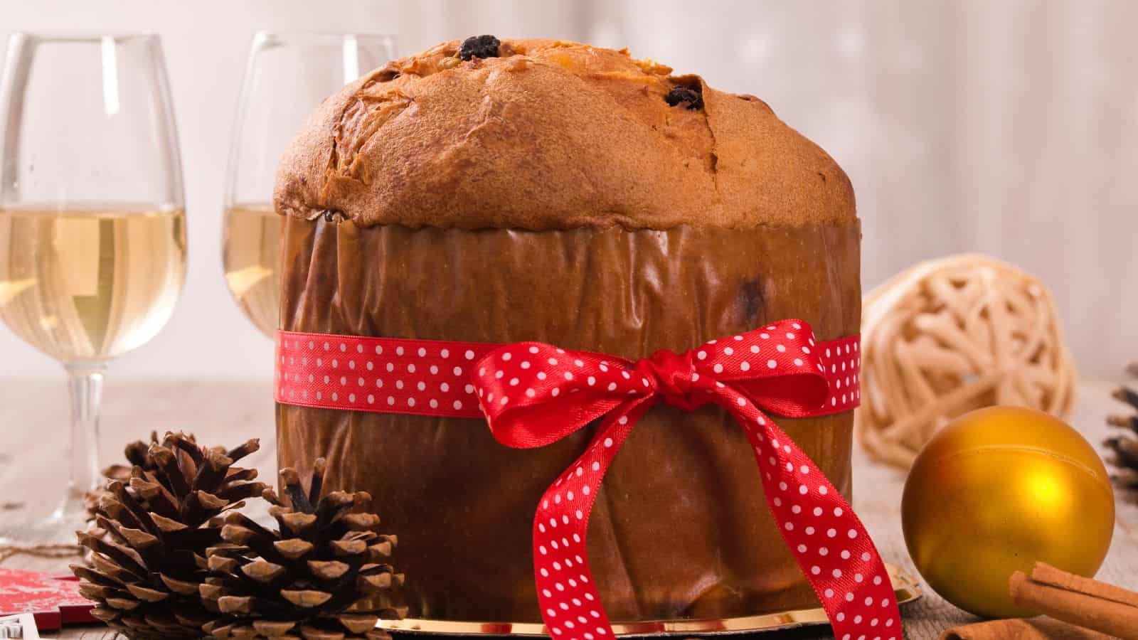 A large panettone wrapped in brown paper and tied with a red ribbon sits on a table with pinecones, a gold ornament, two glasses of white wine, and a decorative ball in the background.