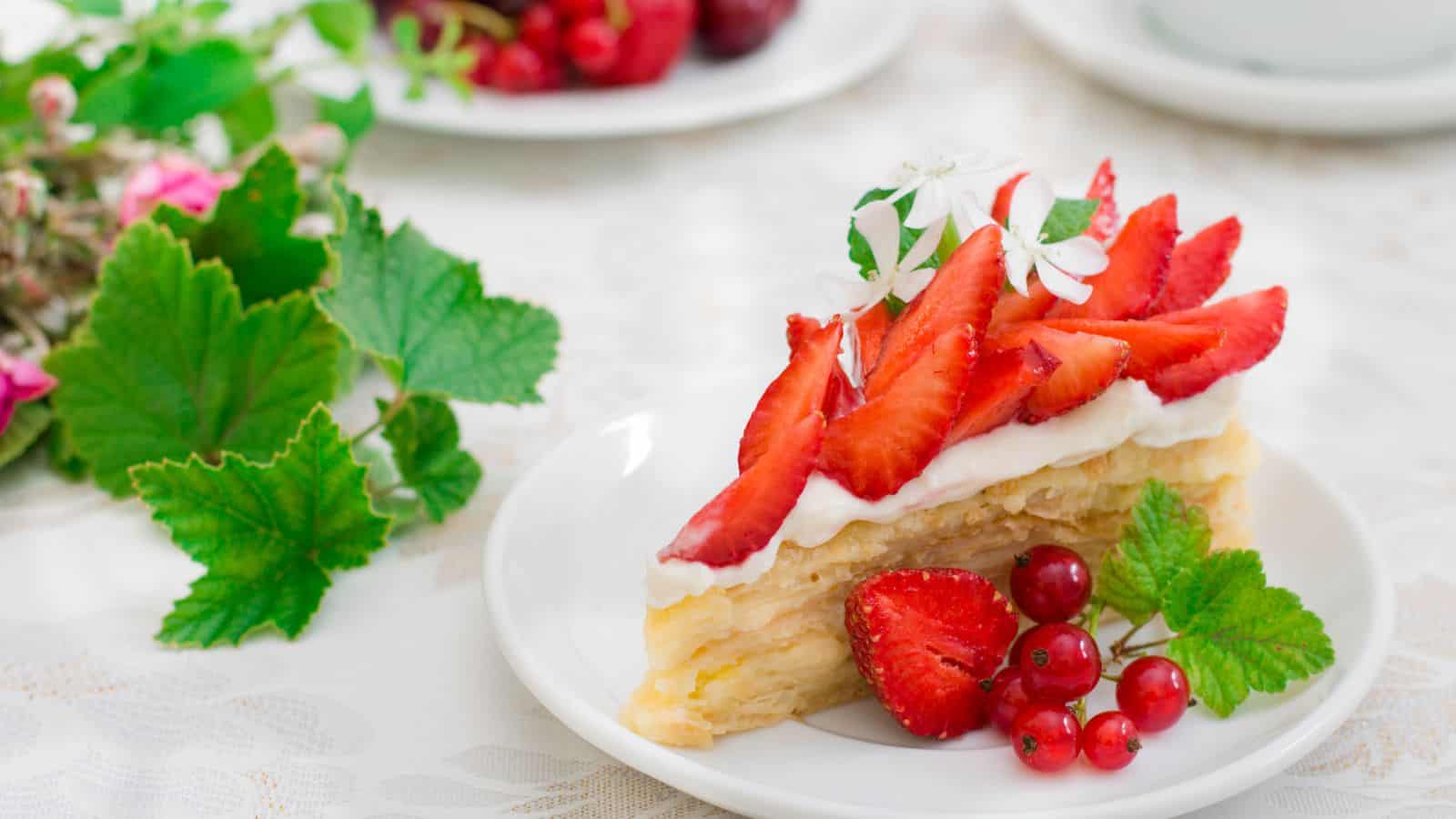 A slice of layered cake with cream, topped with sliced strawberries, red currants, and small white flowers, sits on a white plate. Green leaves and a plate of cherries are visible in the background.