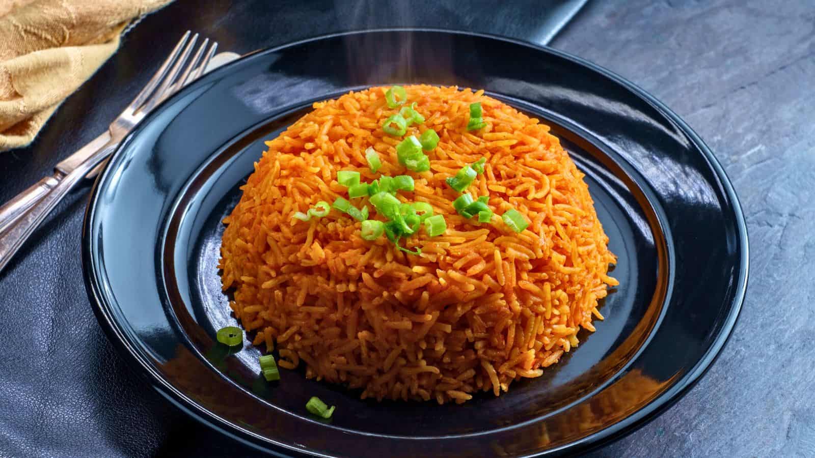 A mound of orange-colored rice garnished with chopped green onions is served on a glossy black plate, with a fork and knife placed beside the plate on a dark textured surface. Steam rises from the rice.