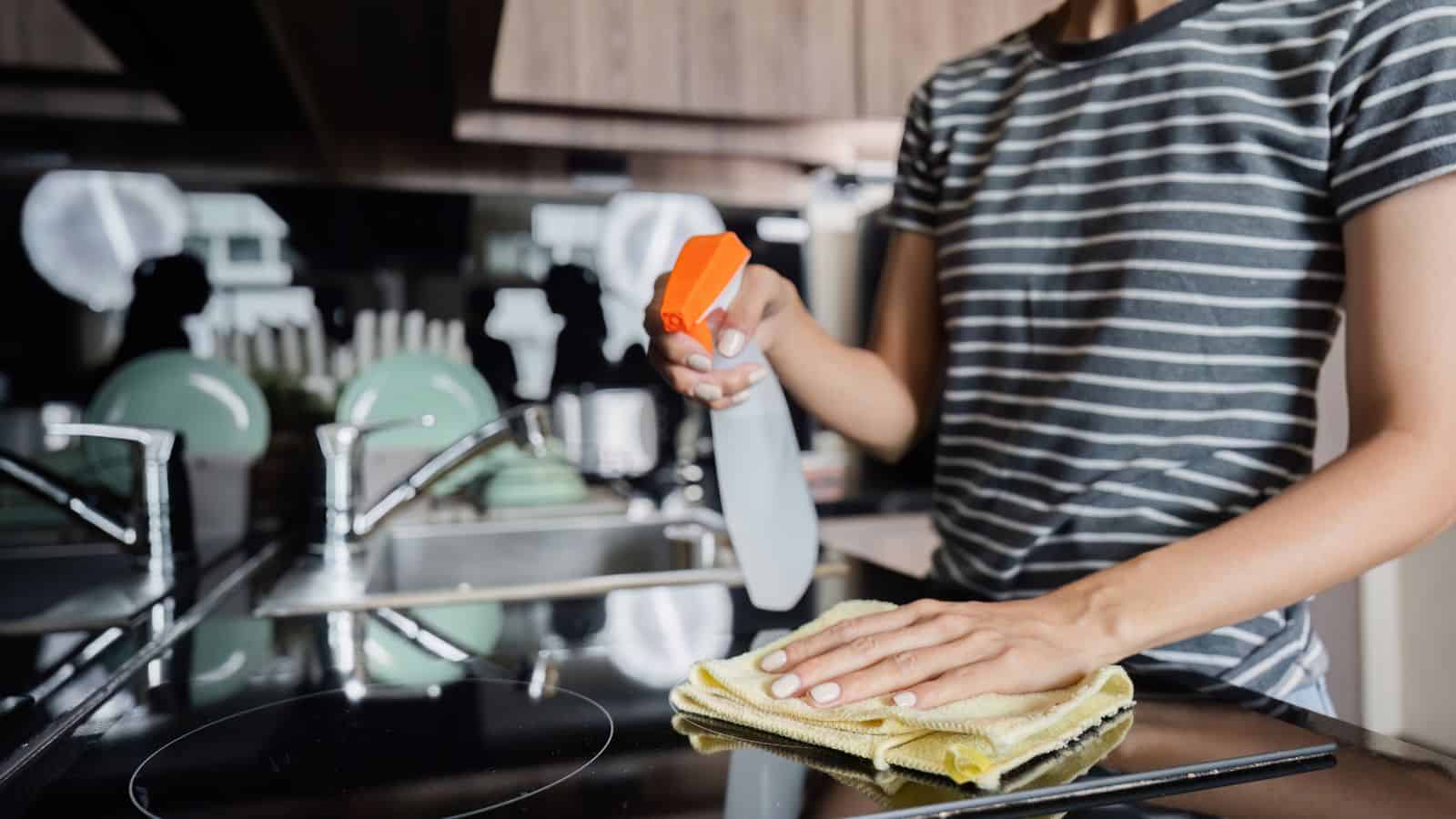 A person wearing a striped shirt uses a yellow cloth and a spray bottle to clean a stove top in a modern kitchen.