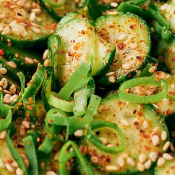Close-up of sliced cucumbers topped with green onions, sesame seeds, and red chili flakes, showing a fresh and seasoned vegetable dish.