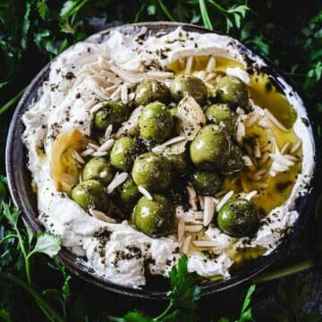 A bowl of Labneh Dip with Warmed Olives, topped with green olives, slivered almonds, olive oil, and sprinkled herbs, surrounded by fresh parsley leaves.