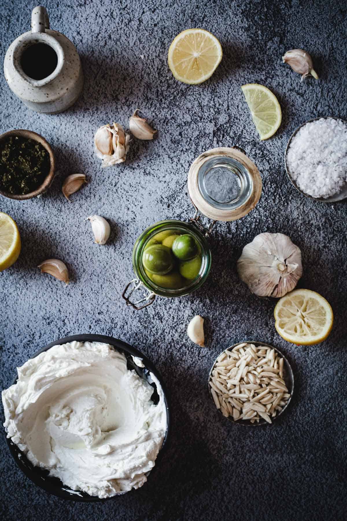 A flat lay of ingredients for Labneh Dip with Warmed Olives on a dark counter, featuring whipped cheese, lemon slices, garlic bulbs and cloves, a jar of olives, slivered almonds, fresh herbs, salt, a small pitcher, and a glass of water.