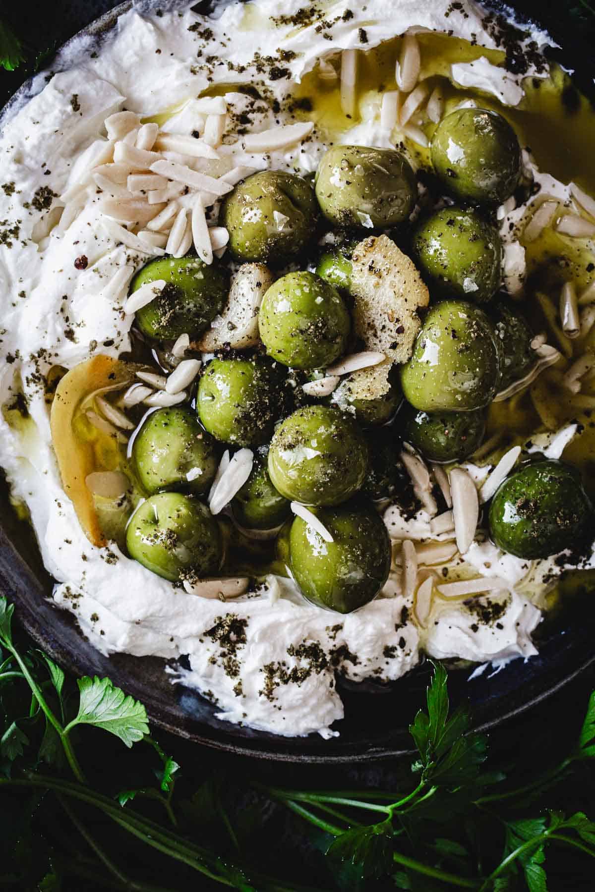 A bowl filled with whipped white cheese, this Labneh Dip is topped with green olives, slivered almonds, olive oil, herbs, and surrounded by fresh parsley leaves.