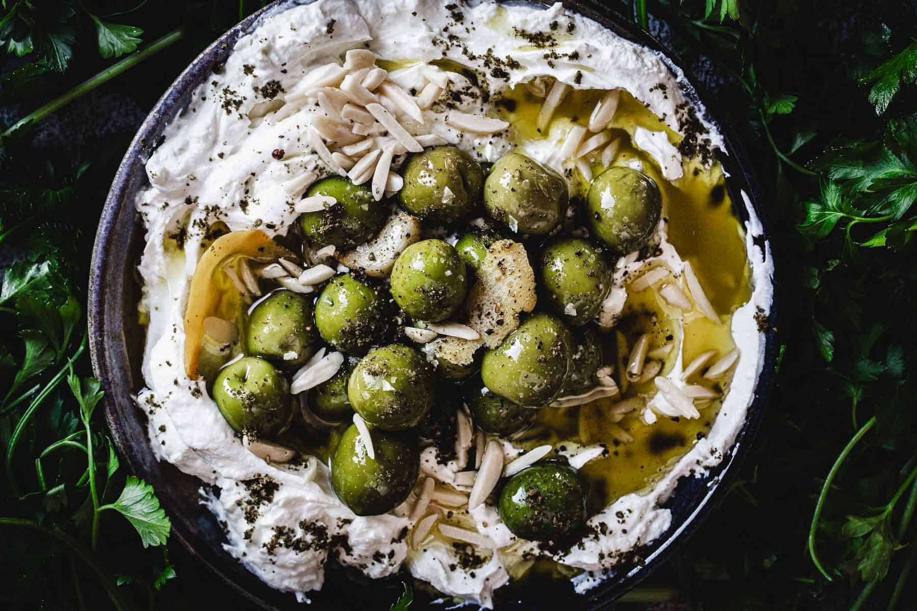 A bowl of whipped labneh dip topped with whole green olives, slivered almonds, a drizzle of olive oil, and a sprinkle of herbs, surrounded by fresh parsley leaves—an inviting Labneh Dip recipe to savor.