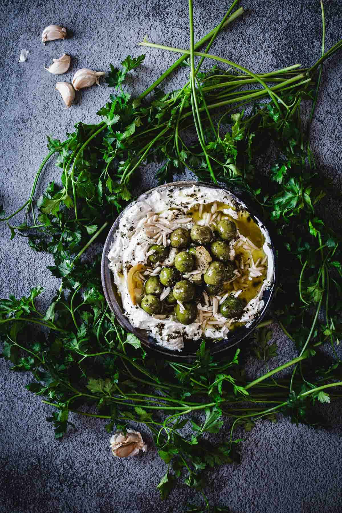 A bowl of creamy Labneh Dip topped with green olives and herbs, surrounded by fresh parsley and garlic cloves on a textured gray surface.