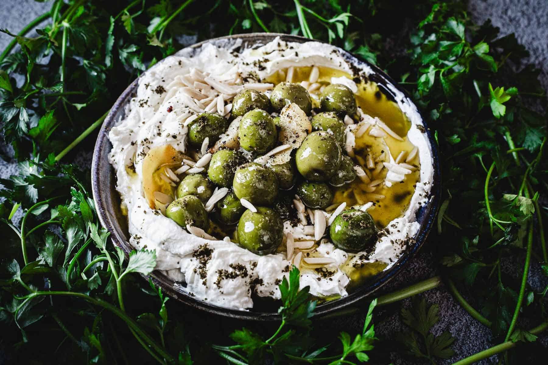 A plate of Labneh Dip topped with green olives, slivered almonds, olive oil, and herbs, surrounded by fresh parsley on a dark surface.