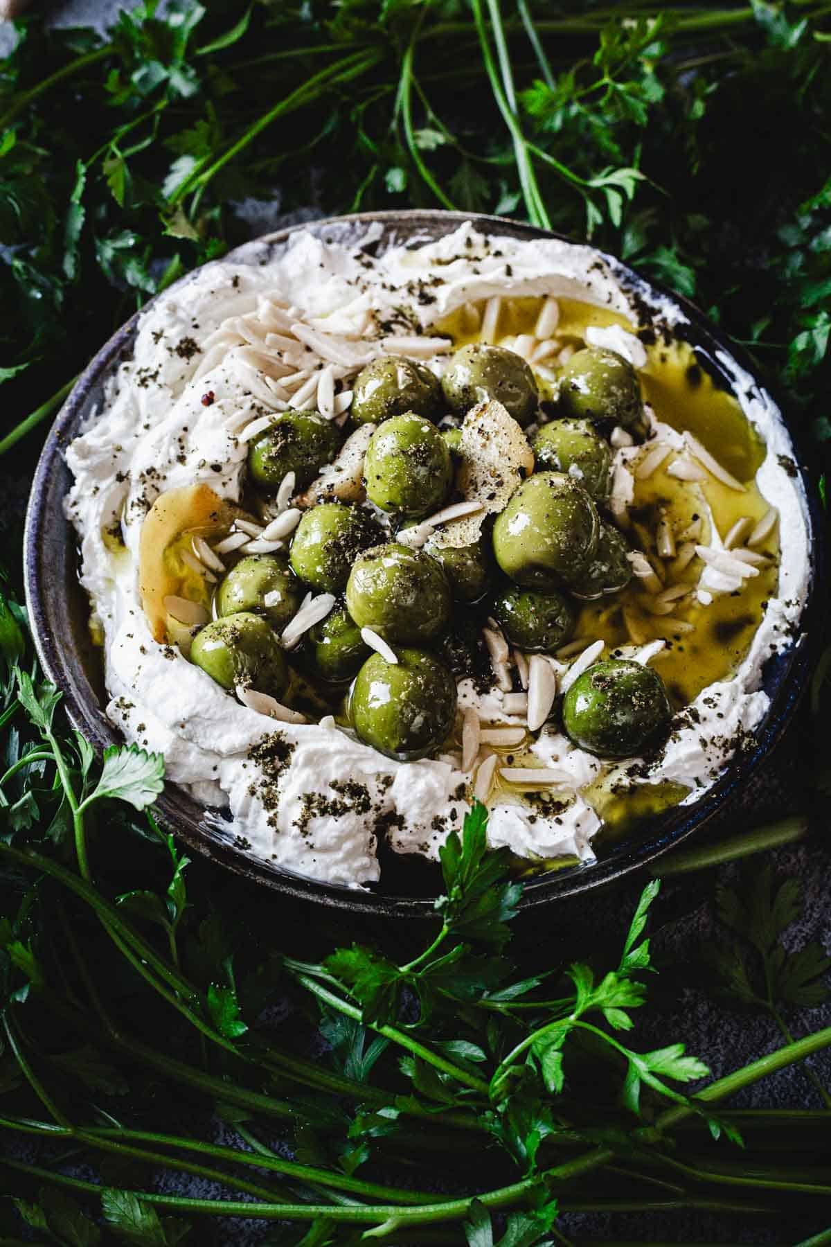 A bowl of whipped feta cheese topped with warmed olives, slivered almonds, olive oil, and herbs, surrounded by fresh parsley sprigs—perfect for dipping or as a unique twist on a classic labneh dip.