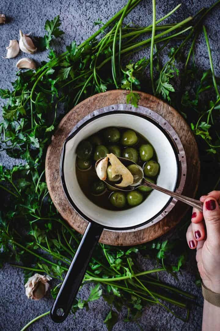 A hand holding a spoon lifts garlic clove from a small pot of warmed olives in brine, set on a wooden board surrounded by fresh parsley and garlic cloves on a gray surface.