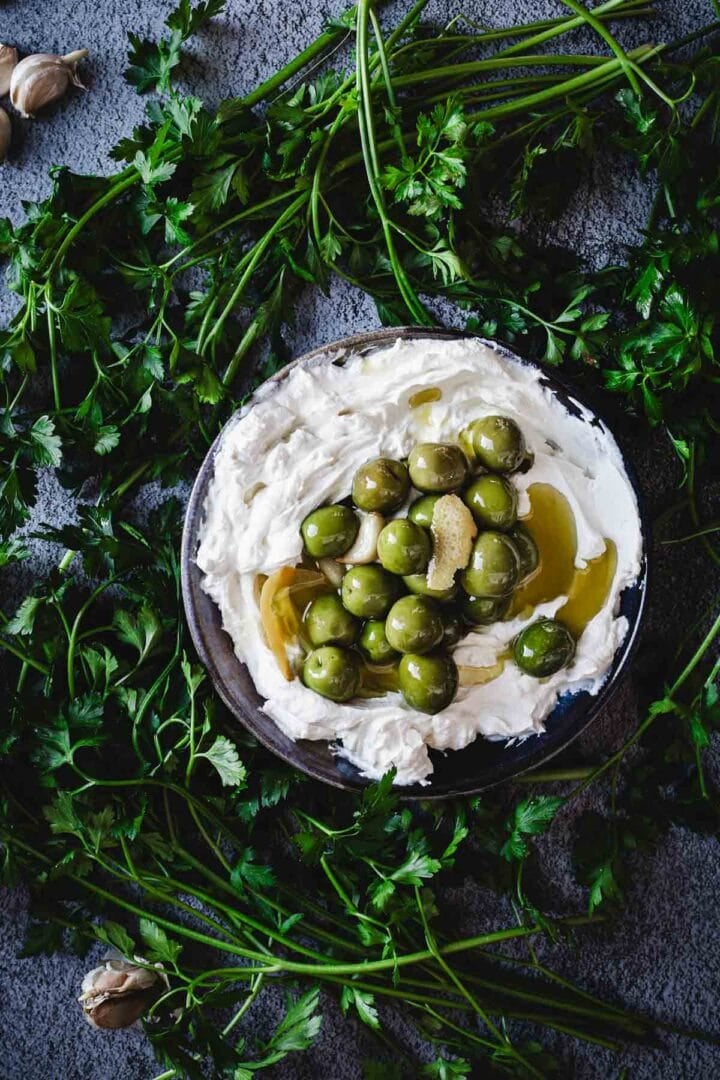 A bowl of whipped ricotta cheese topped with warmed olives and a drizzle of olive oil, surrounded by fresh parsley sprigs on a gray surface—a twist inspired by classic labneh dip.