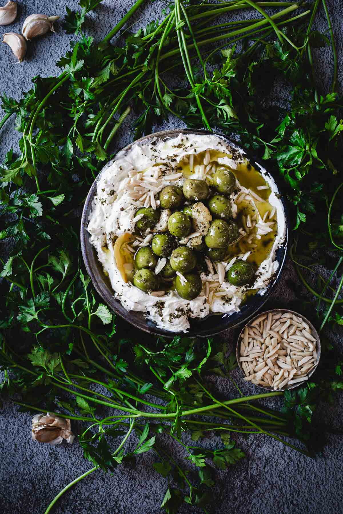 A bowl of whipped feta topped with green olives, slivered almonds, olive oil, and herbs, surrounded by fresh parsley and a small bowl of almonds on a gray surface—perfect inspiration for your next Labneh Dip Recipe. Garlic cloves are also visible.