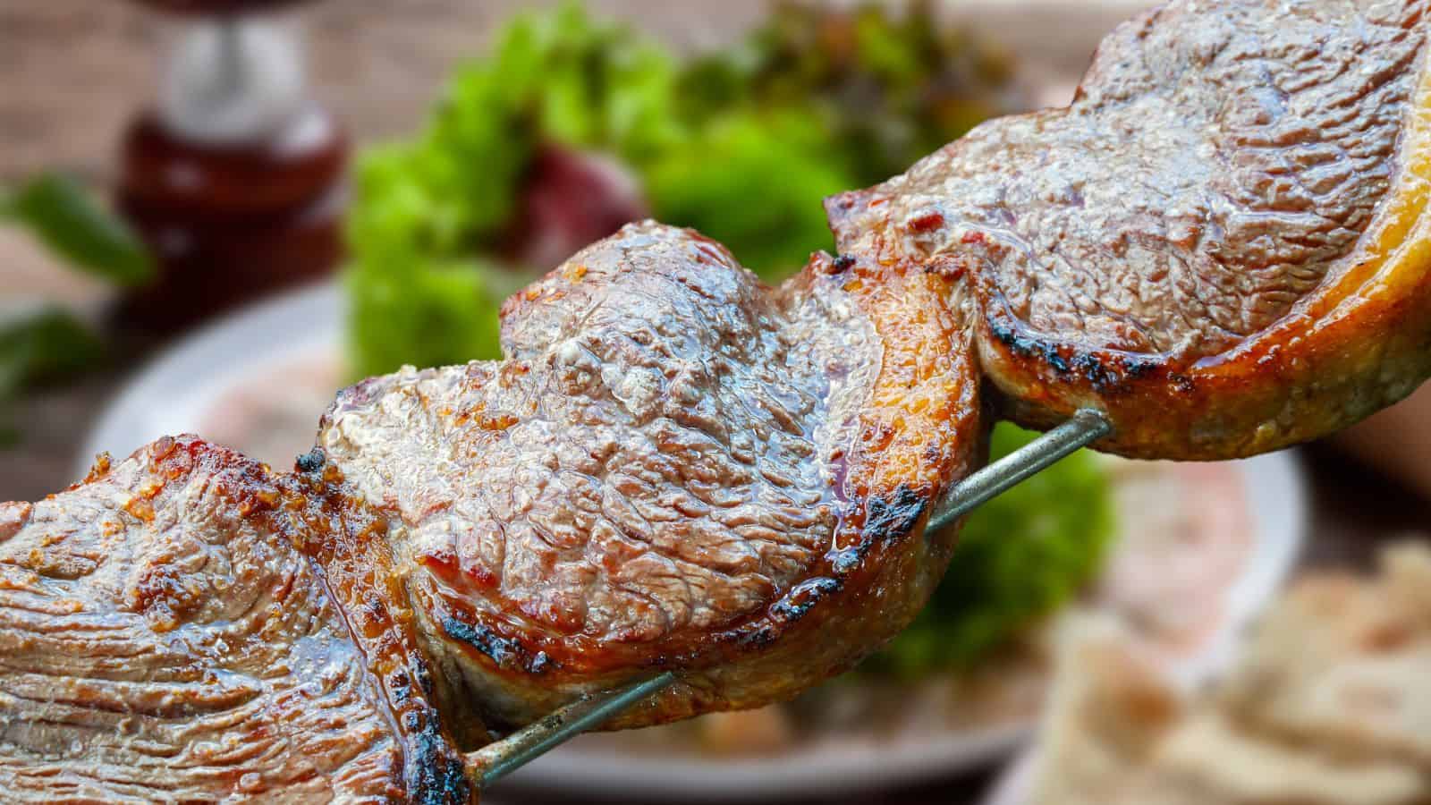 Three pieces of grilled meat on a metal skewer, with a blurred background showing green lettuce and other food items on a table.