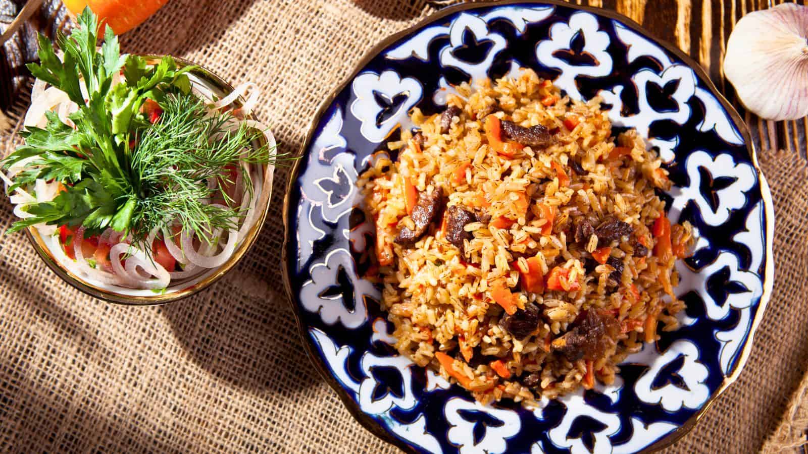 A patterned plate filled with cooked rice, chunks of meat, and vegetables sits on a burlap surface. Next to it is a bowl of sliced onions, tomatoes, and fresh herbs. Garlic and a fork are visible nearby.