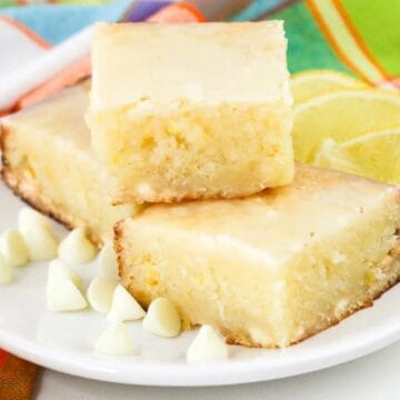 Two glazed lemon brownies are stacked on a white plate, surrounded by white chocolate chips and slices of lemon. A colorful cloth napkin is in the background.