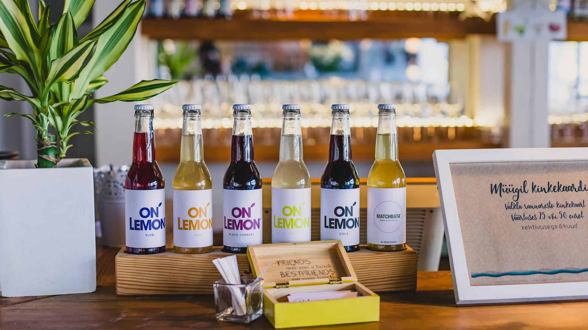 Six "On Lemon" beverage bottles sit on a wooden stand beside a potted plant, business cards, and a sign with foreign text—a reminder to look out for 13 misleading food labels even in stylish displays.