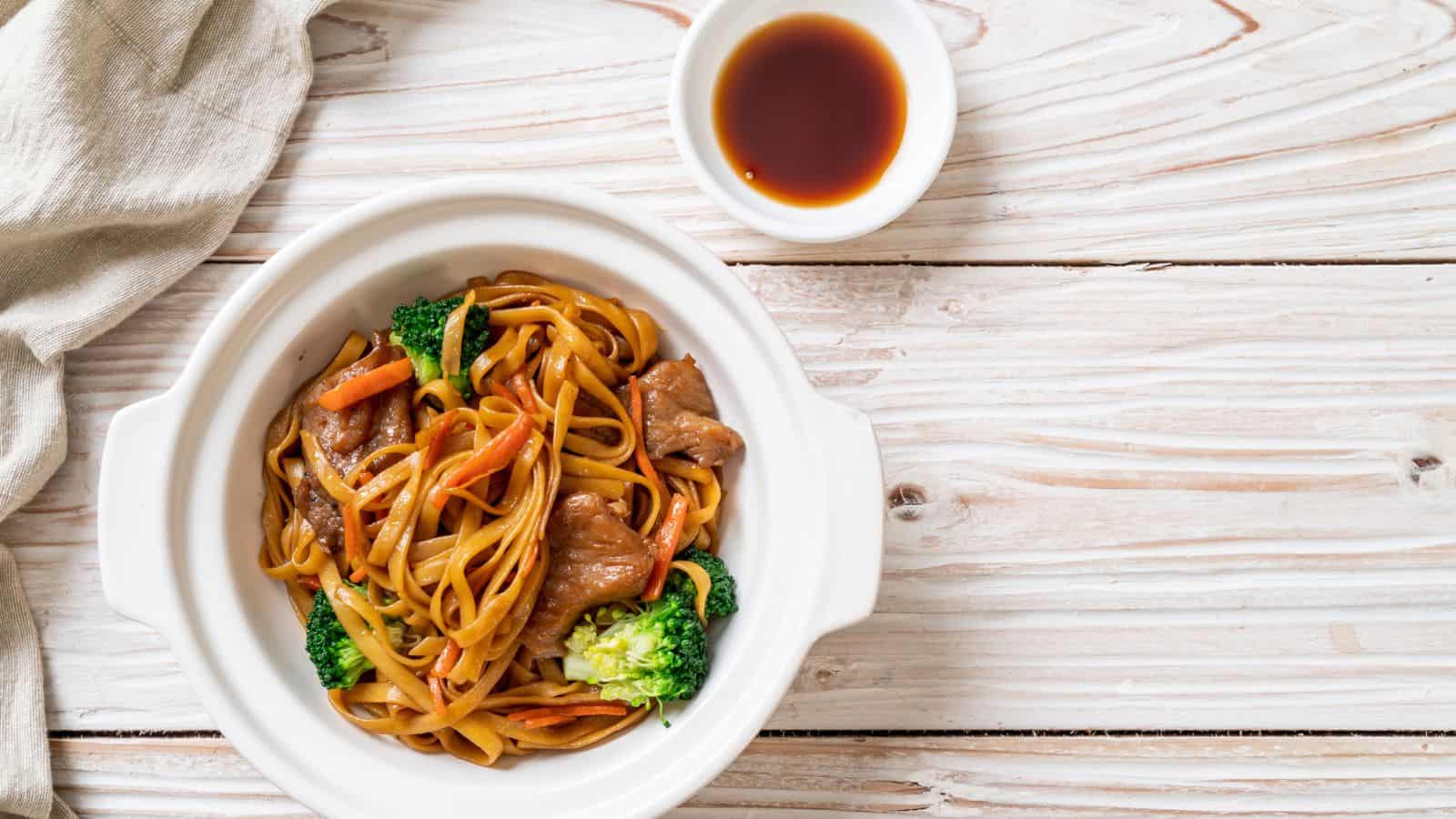 A white bowl filled with stir-fried noodles, beef, broccoli, and carrots sits on a light wooden surface. A small dish of soy sauce and a beige cloth are placed nearby.
